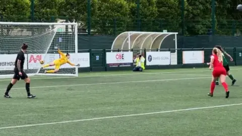 BBC A group of women, one team in red the other in green, playing football - the goal keeper in yellow is diving to their left to save a ball which has gone out of play.
