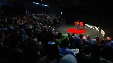 Promote-WIE Nargis on a stage in auditorium surrounded