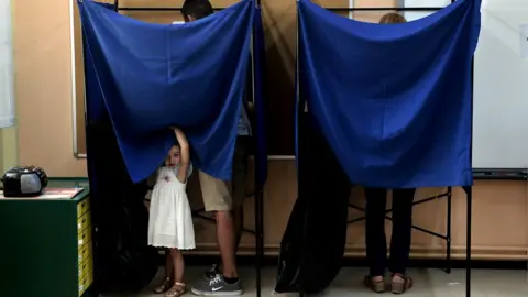 AFP A child peers out from behind the curtain of a polling booth as adults vote in Greek elections
