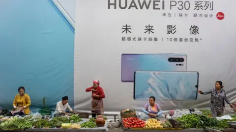 Getty Images Women sell vegetables in front of a billboard advertising smartphones for China's Huawei Technologies Co., on June 1, 2019 in Mangshi, Yunnan Province, southwestern China