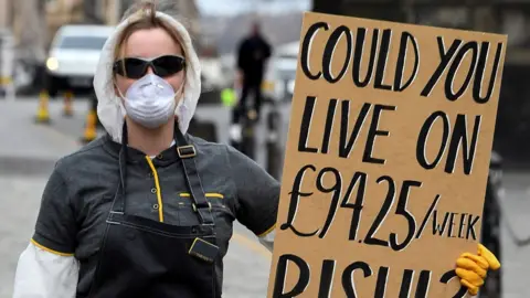AFP A woman wearing a protective face mask and gloves holds a placard complaining about the lack of support for the self-employed and zero hours contract workers, on the Royal Mile in Edinburgh in Scotland on March 23