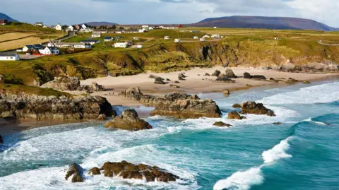 Getty Images Houses in the village of Durness on the north Highland coast. There are sea cliff down to a beach. The sea is a turquoise colour and waves are crashing in on the shore. 
