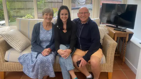 Sofiia Rozhdestvina Allison, with short, grey hair, is on the left and wearing a blue floral dress and cardigan. Soffiia, with long-ish dark hair, is in the centre and has a black top and jeans on and David, with limited hair and wearing glasses, has a blue top and orange shorts. They are seated together in a conservatory on a whicker sofa.