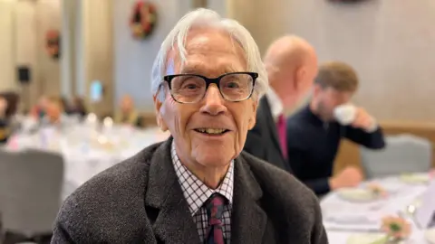 George Carden/BBC Terry Sadler smiling at the camera. He is wearing a suit and tie, glasses and has grey hair. In the background, you can see tables with formal place settings. 