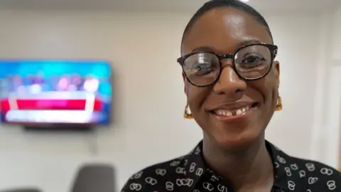 Nicole Lawal smiles and looks directly at the camera. She has dark glasses on and amber earrings and is wearing a black shirt with a white pattern. There is a TV screen in the background.