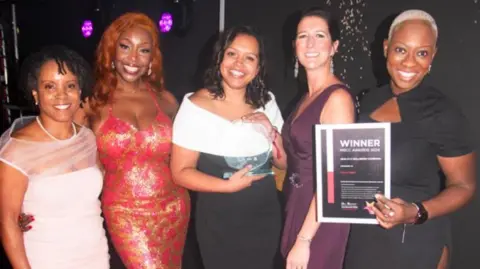 MBCC Five women posing against a black backdrop, with the woman on the right hand side holding up a paper that says winner. The women are dressed in ballgowns 