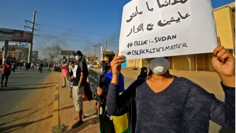 Getty Images A Sudanese protester holds up a sign reading in Arabic "our martyrs are not dead, they are alive with the revolutionaries" along with the English slogans "#BLUEforSUDAN" and "#BLACKLIVESMATTER", as demonstrators mark the first anniversary of a raid on an anti-government sit-in and some demonstrate in support of US protesters over the death of George Floyd, in the Riyadh district in the east of the capital Khartoum on June 3, 2020