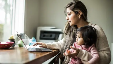Getty Images Woman holding a toddler while using a laptop.