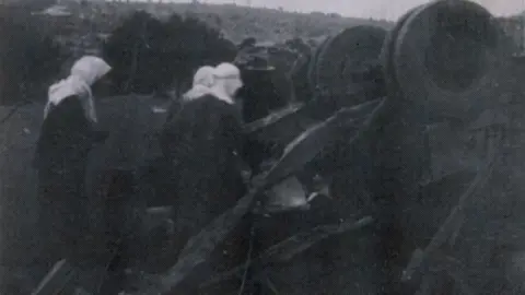 Harry Arrigonie/Edward Gaskell A black and white photo of women surrounding upturned remnants of a bus