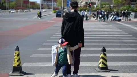 Getty Images A man leading a face mask-clad child waits to cross a street in Beijing on April 11, 2020, amid the COVID-19 coronavirus pandemic