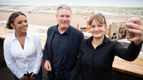 PA Media Sir Keir Starmer, the leader of the Labour Party, takes a selfie on the Worthing sea front with two young women. Sir Keir, a white man in his 60s with greying hair, wears a dark blue shirt and suit jacket. He stands between two young women, one of whom is holding her iPhone aloft to pose for the selfie she is taking. The woman on Sir Keir's right smiles at the phone and wears a white shirt and has her dark curly hair tied back. On Sir Keir's right, the young woman wears a black short and has her blonde hair tied back. Behind them is the sandy beach and sea which looks green on an overcast day.