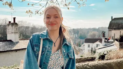 Jodie Powell Jodie Powell is an 18-year-old white woman, she wears her long blonde hair tied back with her pony tail pulled over her left shoulder. She wears a denim jacket over a floral dress with a canvas tote bag over her right shoulder and a charm necklace on a black string. She is smiling at the camera and is photographed outside, with the roofs of houses and chimneys visible behind her as well as a line of trees against a pale blue sky