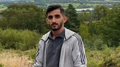 Rizwan Baig Rizwan Baig pictured on a rural hill surrounded by greenery on an overcast day. Rizwan is an Asian man in his 20s and has short dark hair and a cropped beard. He is wearing a light grey Adidas tracksuit jacket over a dark grey polo shirt. He is looking at the camera with a slight smile