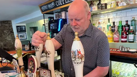 Kate Bradbrook/BBC Malcolm Gray with short grey hair and beard pulling a pint in his pub
