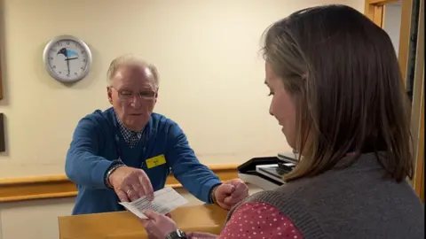 Terry Seston, who is behind a brown desk, hands over a small piece of paper to a woman with shoulder-length brown hair, who is wearing a red polka-dot shirt and grey sleeveless cardigan