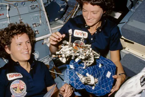 NASA Astronauts Kathryn D. Sullivan, left, and Sally K. Ride display a "bag of worms." The "bag" is a sleep restraint and the majority of the "worms" are springs and clips used with the sleep restraint in its normal application