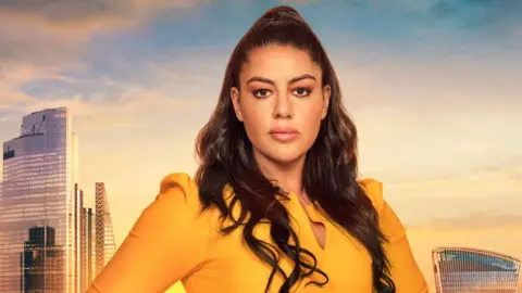 BBC/Naked/Ray Burmiston Picture of Nadia Suliaman in a mustard yellow top. She stands infront of a green screen of London. She looks blankly into the camera and has brown eyes and dark brown hair. 