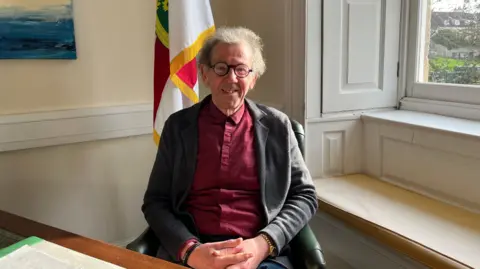 A man with purple glasses, red shirt and grey cardigan smiles at the camera. The flag of Alderney is behind him