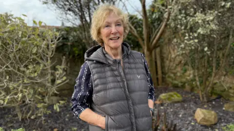 BBC A woman with blond hair looking at the camera. She is wearing a striped navy blue shirt with a black gillet. Behind her is a garden. 