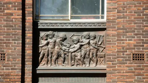 Historic England A bronze plaque set into the wall of the building, showing six child figure working on parts of a bicycle