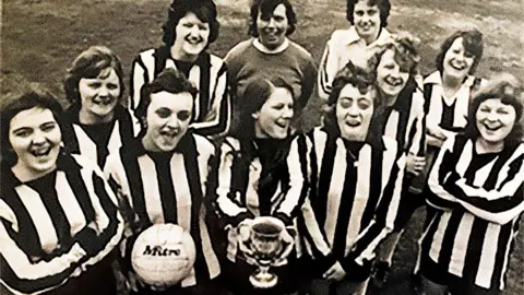 Wolves Women A black and white image of a 11 women in football uniforms with black and white vertical stripes. They are all smiling. In the centre of the group one of the women is holding a trophy, while the woman next to her is holding a football