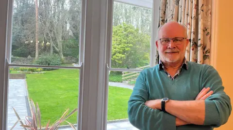 A man standing with his arms folded beside a window smiling at the camera