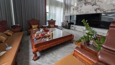 BBC/Tony Han A two-floor-high living room with floor-to-ceiling windows and curtains. The space is clad in marble, and furnished with a coffee table and sofas carved out of wood in a rich red-brown colour. It also features a large flatscreen television.