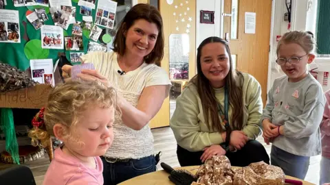 BBC A group of women and children sitting together at a nursery in Somerset. A little girl with curly blonde hair is having it styled by a brunette woman