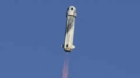 Getty Images Blue Origin’s New Shepard rocket lifts off vertically with capsule attached, emitting a bright exhaust plume against a clear blue sky.