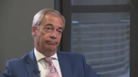 Nigel Farage, sitting down wearing a dark blue jacket, white shirt and pink tie