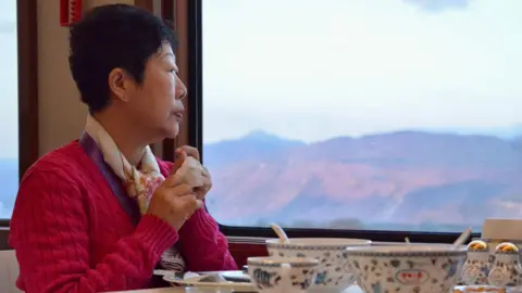 BBC/Rachel Yu A woman with black, short hair, wearing a red jumper and scarf around her neck is sitting at a train table where there are traditionally-ornamented bowls, a teacup and salt and pepper pots, looks out of the window at a reddish mountainside and pale blue sky.