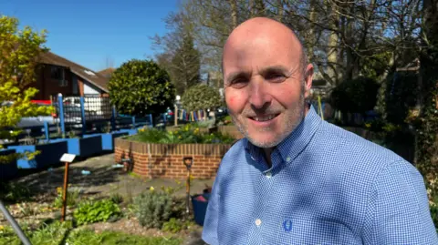 Keith Sinclair sitting outside on a sunny day wearing a light blue hecked long-sleeved shirt. Behind him but not in focus is a planned garden with small walls and planters
