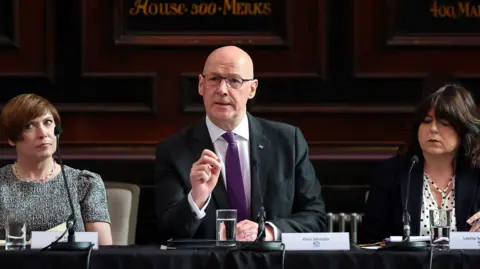 PA Media John Swinney - wearing a dark suit with purple tie - sits behind a desk, with a microphone and glass of water in front of him. He has his hand raised. Two women sit next to him listening - one is wearing a grey top and the other with a white top and black jacket.