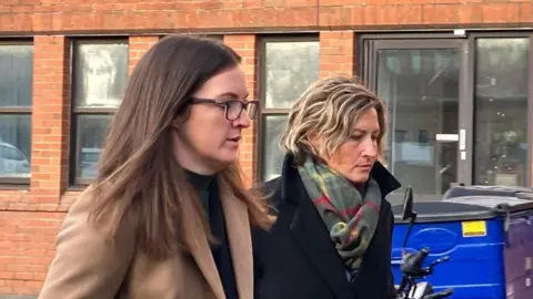 Two women walking alongside eachother past a red brick building. One is wearing a camel coat, glasses and has dark brown hair. The other, Nerys Lloyd, is wearing a dark coat and a multicoloured scarf.
