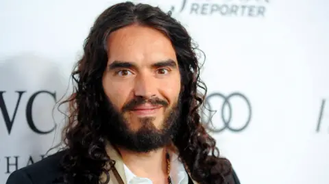 Reuters Russell Brand has long curly hair, a beard and is wearing a white shirt and dark jacket. He stares intently at the camera and smiles