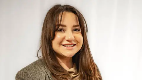 Emily Bateman Photography Danielle Cox standing in a room by a wall with long dark hair, wearing a jacket and beige top. She is smiling and looking straight at the camera. 