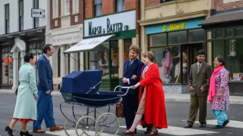 Black Country Living Museum Men and women dressed in 1950s attire cross a road at the museum. One couple is pushing a pram with a South Asian couple walking behind them and another couple walking towards them. 