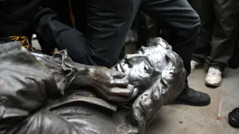 PA Media The Edward Colston statue on the floor after being toppled by protesters in Bristol. The metal figure depicts Colston with his chin resting in his hand. A protester wearing black jeans is kneeling on the statue's neck. 