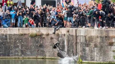 Reuters A crowd of protesters at Bristol Harbour as the Edward Colston statue is dumped into the water. Many people are wearing facemasks. Most of them are taking photos on their phones or cameras. The statue is half in the water and there is a splash where it enters. 