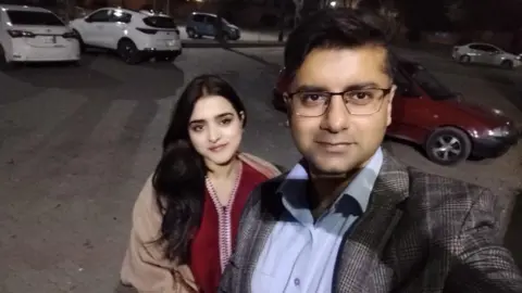Sheharyar Shahnawaz Haleema pictured standing slightly behind Sheharyar in a selfie he is taking of the two in a car park in the dark. She has long black hair and wears a red tunic and beige coat. He wears glasses, a grey checked blazer and a blue shirt