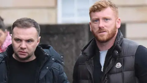 Getty Images Eddie Lyons Jnr, who has short brown hair and is wearing a dark blue raincoat and black t-shirt, next to Ross Monaghan, who has curly ginger hair and a bear, and is wearing a black puffer jacket, a black zipper underneath and a white t-shirt. 