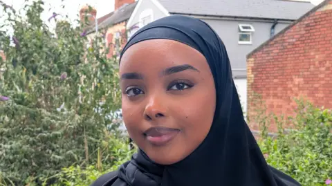 BBC Munirah, a young Welsh-Somali woman stands on a street in Grangetown, Cardiff with trees and houses behind her. She's looking at the camera, smiling and wearing a black headscarf. 
