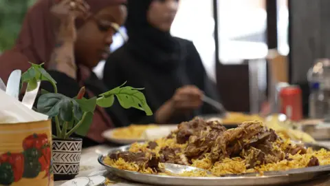 In the foreground is a large plate of lamb and spiced rice with a large serving spoon. In the background two young Somali women are enjoying their meal. 