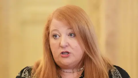 PA Media Naomi Long has medium length ginger hair in a side parting with blue eyes. She is mid speech and her background is yellow and blurred out. She's wearing red lipstick, a silver choker necklace and a black and white patterned top.