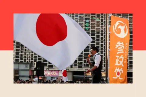 Reuters Japan's Sanseito party leader Sohei Kamiya delivers a speech during the party’s rally - large flag in the foreground and Sohei wears a white shirt and holds a microphone