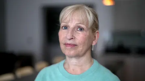 Brijesh Patel / BBC Caroline looks into the camera - she has short, light-colored hair and is wearing a light blue sweater. The setting appears to be indoors with low lighting. In the background, indistinct furniture is visible, contributing to a subdued atmosphere. An orange light source glows in the upper right corner, casting a warm hue over part of the scene.