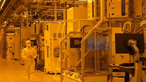 Getty Images A staff member in hazesuit walks in the cleanroom of a semiconductor manufacturing facility at GlobalFoundries in Singapore on September 12, 2023. He is surrounded by large machines that make chips. 