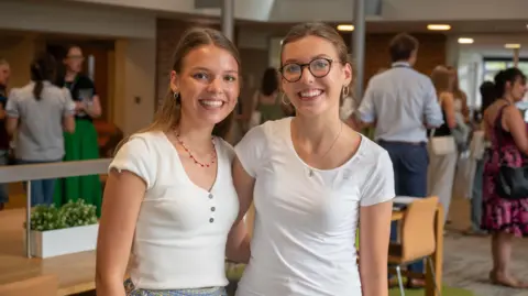 Headington Rye Oxford Florence and Alice Bolton stand side by side looking at the camera and grinning widely. They both have dark blonde hair and are wearing white shirt sleeved t shirts. In the background are groups of people gathered inside the college talking.