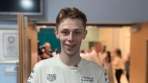 Harry has short blonde hair and smiles at the camera as he wears a white t-shirt with branding on it. Behind him the background is blurred - the open doors of a school assembly hall can be seen with people milling around inside it.