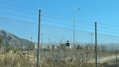 A barbed wire fence and behind it some buildings and a mountain